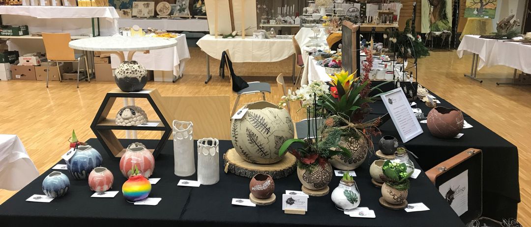 Ein Raum gefüllt mit Keramikvasen zur Ausstellung, einige mit Blumen, auf einem schwarzen Tisch angeordnet. Dahinter Tische mit verschiedenen Gegenständen, eine Treppe und Kunstwerke.