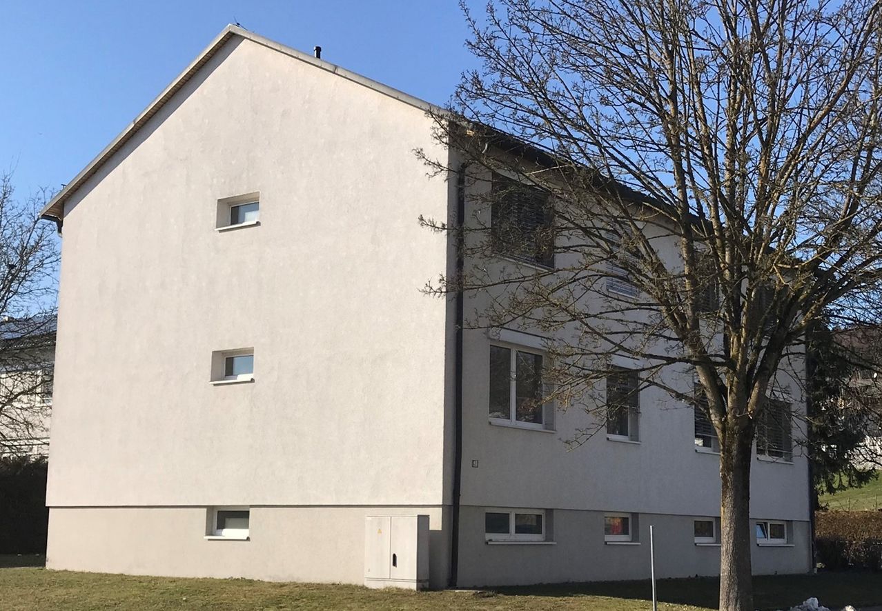 Ein dreistöckiges Gebäude mit einem Schornstein auf dem Dach und mehreren Fenstern. Davor steht ein kleiner Baum.