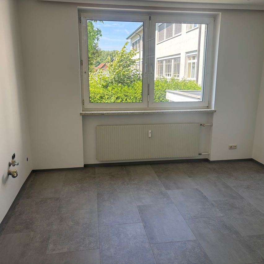 Ein leerer Raum mit weißen Wänden und grauen Fliesen auf dem Boden. Es gibt ein Fenster mit weißem Rahmen, und ein weißer Heizkörper ist an der Wand unter dem Fenster montiert.