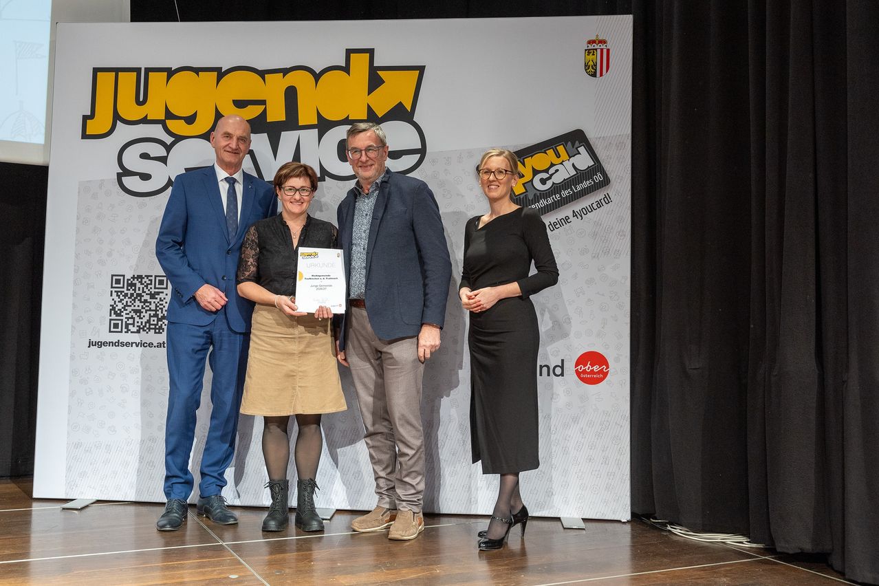 Drei Personen stehen auf einer Bühne mit einem Hintergrund mit Text. Die Frau in der Mitte hält ein Zertifikat, während die beiden Männer neben ihr stehen. Der Hintergrund hat Logos und Text.