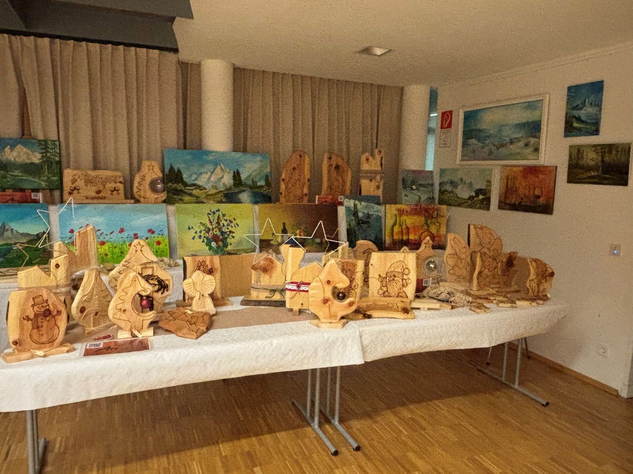 Ein Tisch zeigt verschiedene Holzkunstwerke, darunter Schnitzereien und Gemälde, in einem Raum mit Holzböden und Vorhängen.