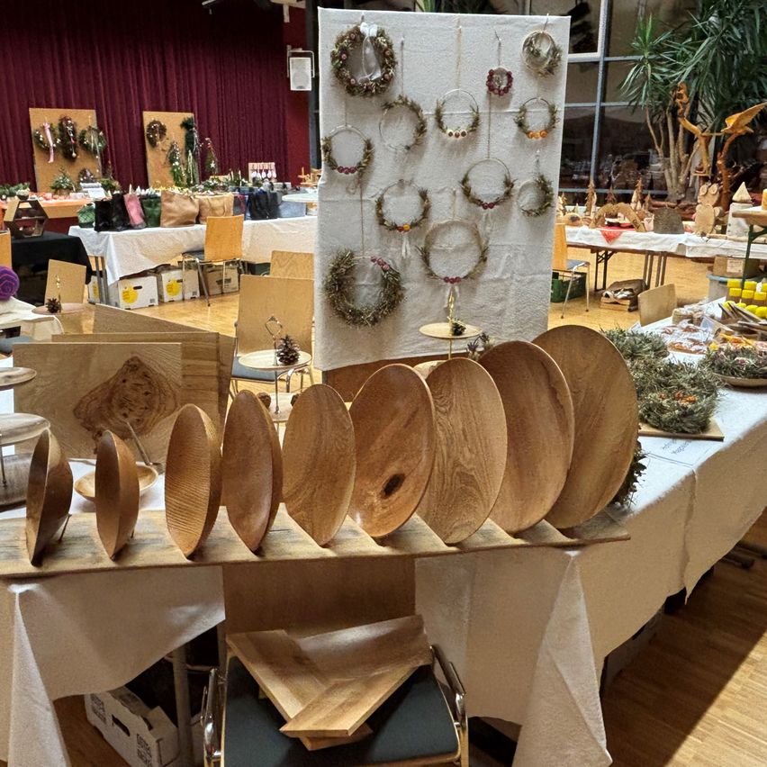 Ein Raum voller Holzarbeiten, ein Holztisch im Vordergrund mit einer Reihe von Holztellern und dahinter sind verschiedene Handwerksarbeiten auf Tischen und an der Wand ausgestellt.