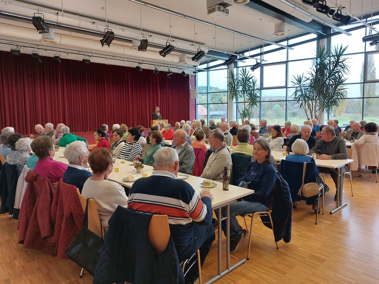 Eine Gruppe von Menschen sitzt um Tische herum in einem Saal. An den Seiten stehen Topfpflanzen. Eine Person steht an einem Rednerpult, möglicherweise im Sprechen.