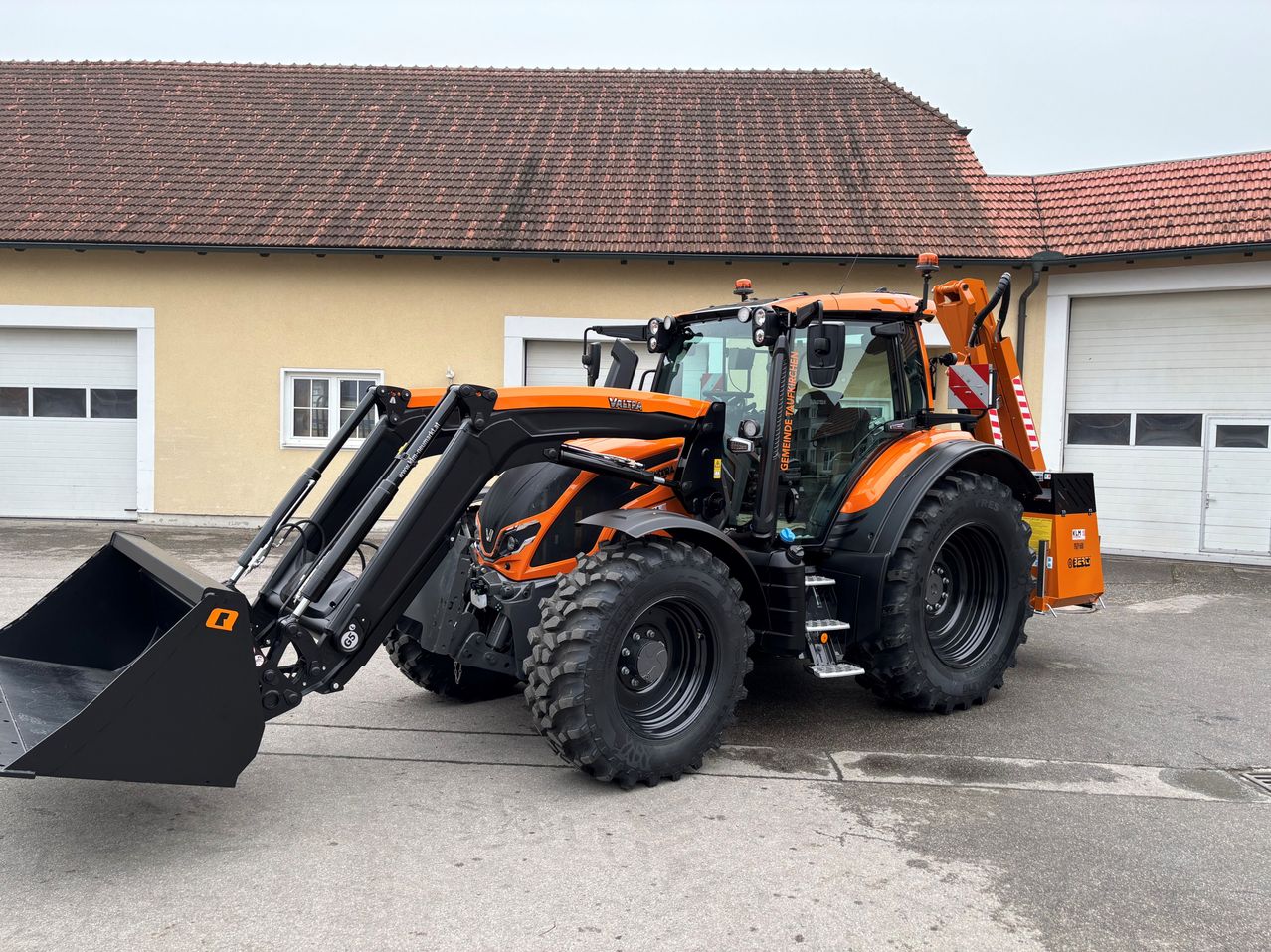 Ein orangefarbener und schwarzer Traktor steht vor einem gelben Gebäude mit einem roten Dach. Der Traktor hat eine große Anbauvorrichtung vorne.