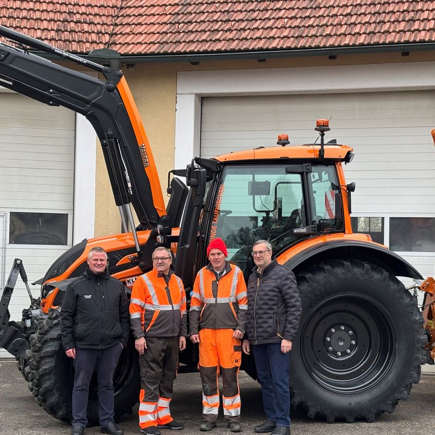 Vier Männer stehen vor einem großen orangefarbenen Traktor. Drei tragen Warnwesten in Orange, einer trägt einen schwarzen Mantel. Sie scheinen für ein Foto zu lächeln.