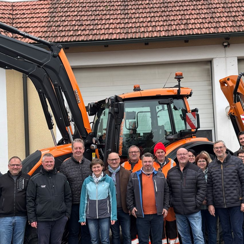 Eine Gruppe von Menschen, darunter Männer und Frauen, steht vor einem orangefarbenen Traktor mit einem Gebäude im Hintergrund.