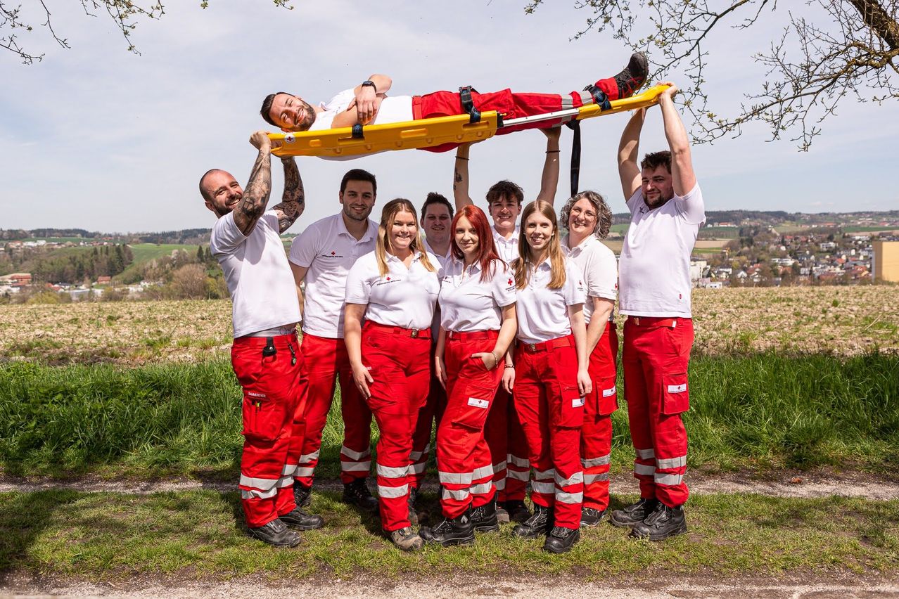 Eine Gruppe von Menschen in roten und weißen Uniformen posiert mit einem Mann auf einer Trage im Freien.