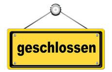 Ein gelbes Schild mit schwarzen Buchstaben steht für 'Geschlossen'. Es hängt an einem Metallhaken. Das Schild ist auf einem weißen Hintergrund platziert.