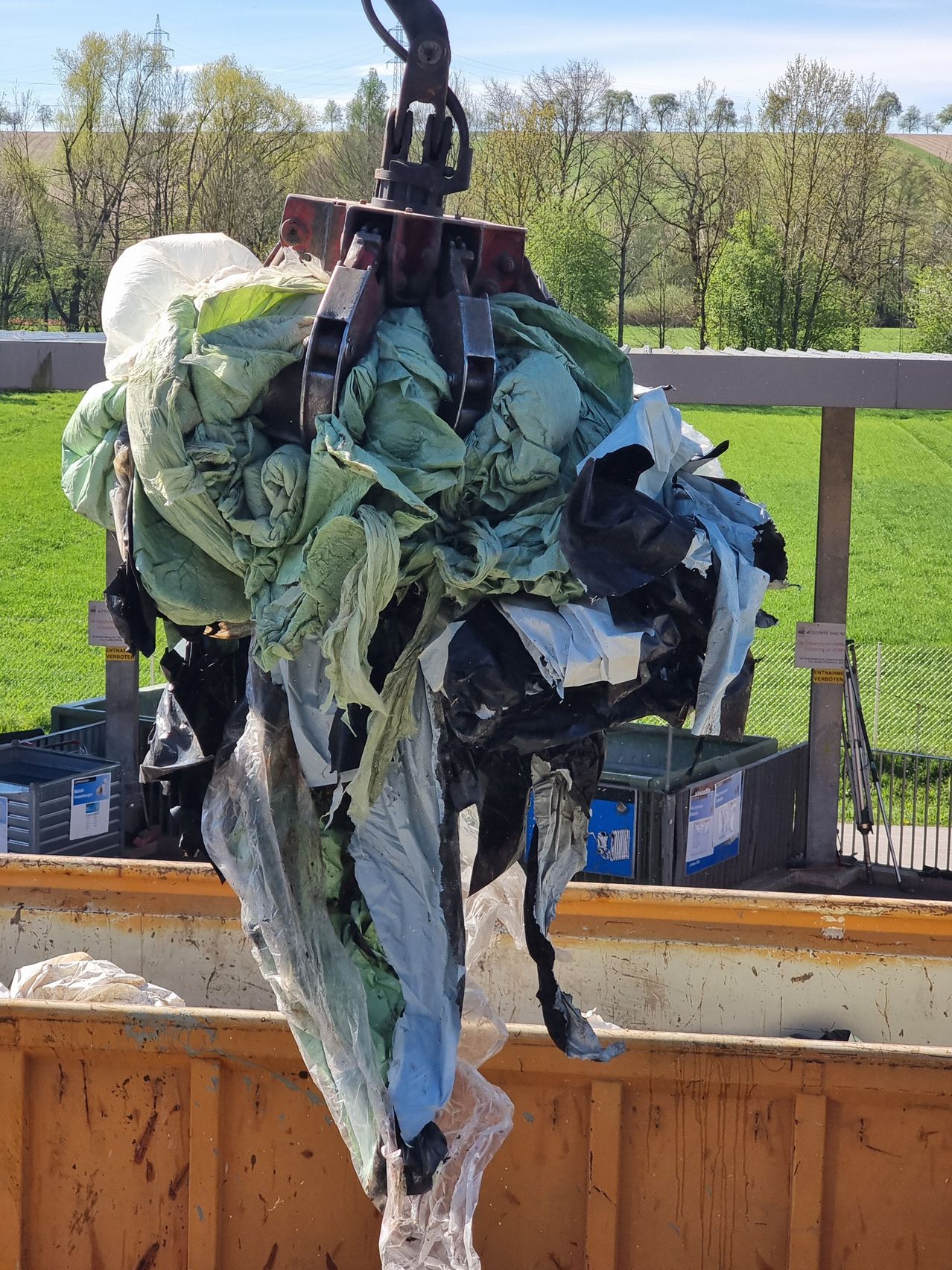 Ein Kran hebt einen Haufen Müllsäcke an, einige zerrissen, vor dem Hintergrund eines grünen Feldes und eines klaren blauen Himmels.