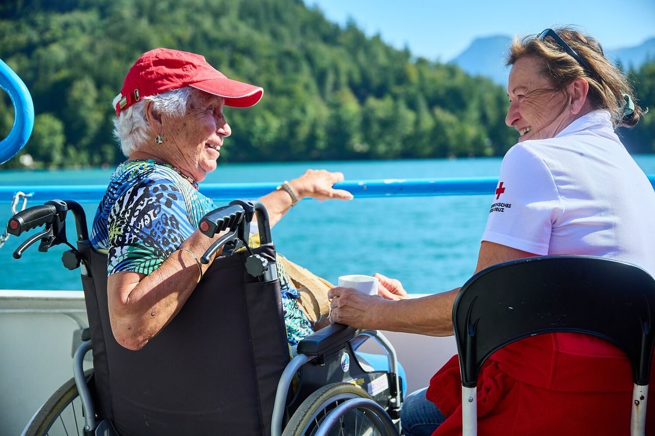 Eine ältere Frau im Rollstuhl ist auf einem Boot, lächelt und gestikuliert, während sie einen Becher hält. Sie ist mit einer anderen Frau in einem weißen Shirt mit rotem Kreuz. Sie sind von einer malerischen Aussicht mit Bäumen und Bergen umgeben.