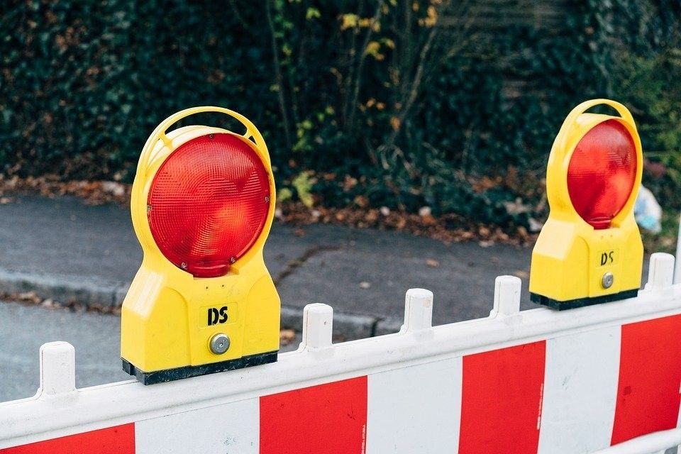 Zwei gelbe Warnleuchten mit roten Linsen sind auf einer weiß-rot gestreiften Barriere auf einem Bürgersteig montiert.