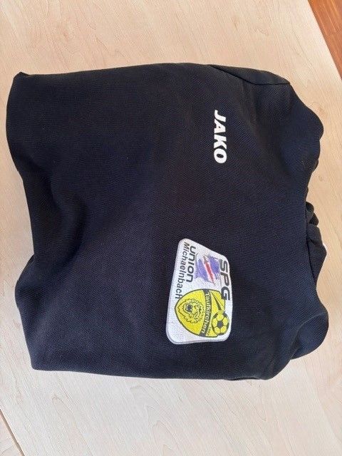 Eine schwarze Sporttasche mit dem Wort 'Jako' darauf gedruckt. Sie hat ein weißes Aufnäher mit einem Fußball und dem Text 'Union Michaelbach'.