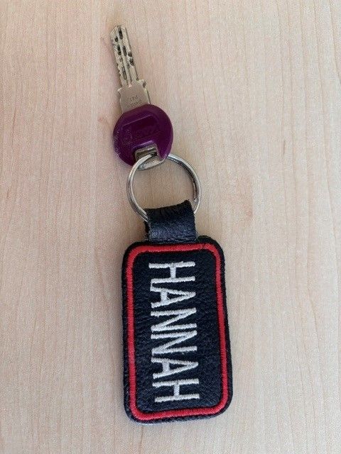 Ein Schlüsselanhänger mit einem Lederschild, auf dem 'HANNAH' steht. Der Schlüssel ist silbern mit einer lila Kappe und einem schwarzen Lederband. Der Anhänger hat einen roten Rand und weiße Nähte.