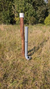 Ein grauer Rohr ist an einem Holzpfahl in einem Grasfeld mit Bäumen im Hintergrund befestigt.