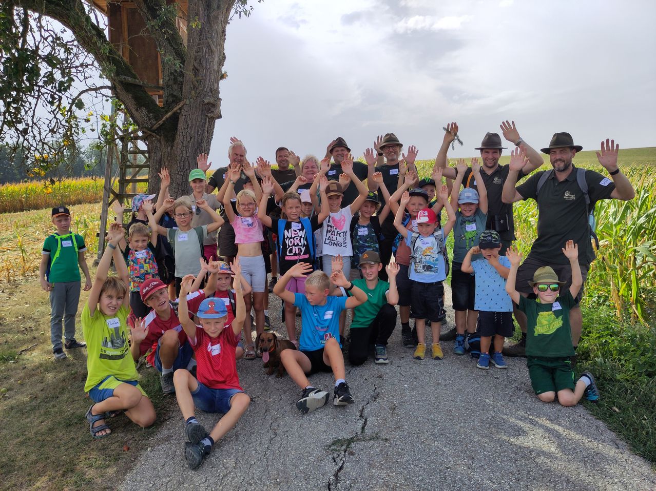 Eine Gruppe von Kindern und Erwachsenen posiert für ein Foto auf einem Feld, einige tragen Hüte und Brillen, mit einem Baumhaus im Hintergrund.
