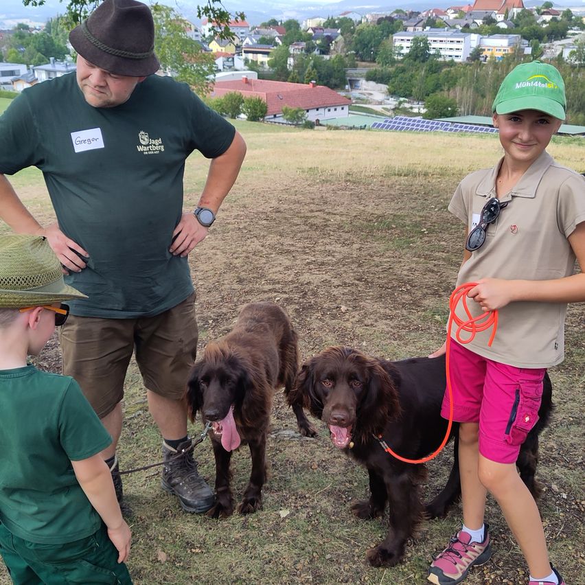 Drei Menschen und zwei Hunde stehen auf einem Feld. Ein Mann und ein Mädchen lächeln, während ein Junge zur Kamera schaut. Der Mann trägt einen Hut und ein grünes Shirt. Das Mädchen trägt einen grünen Hut und pinke Shorts. Der Junge trägt ein grünes Shirt und einen Hut.