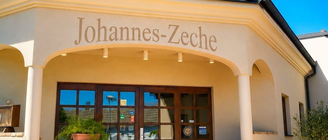Johannes-Zeche-Gebäude mit einer Ziegelfassade, einem Bogeneingang, hölzernen Türen und einem Fass mit einer Pflanze auf den Stufen. Das Schild lautet Johannes-Zeche und Herzlich Willkommen.