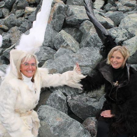 Zwei Frauen in Pelzmänteln posieren zwischen großen Felsen und Eis. Die Frau auf der linken Seite trägt einen weißen Pelzmantel und eine Brille, während die Frau auf der rechten Seite einen schwarzen Pelzmantel trägt.