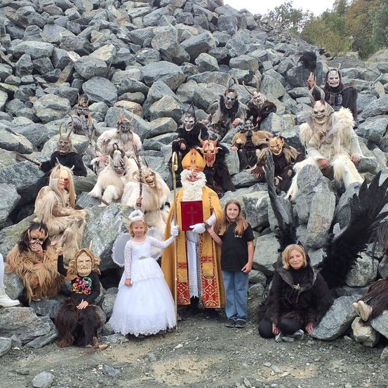 Eine Gruppe von Menschen in Kostümen posiert für ein Bild mit einem verschneiten Hintergrund, einige sitzen auf Felsen und einer ist als Engel gekleidet.