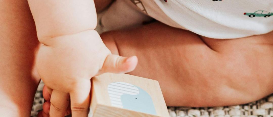 Ein Baby, das ein weißes Strampler mit Auto-Prints trägt, sitzt auf dem Boden und hält einen Holzblock mit einer Wal-Illustration.