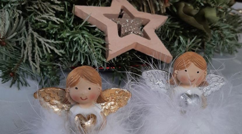 Zwei goldene Engelfiguren mit weißen Federn stehen dicht beieinander auf einer weißen Oberfläche. Dahinter befindet sich ein Weihnachtsbaum mit einem Holzstern.