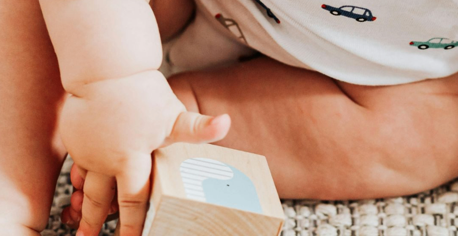 Ein Baby sitzt auf dem Boden und hält einen Holzblock mit einem Wal-Design. Die Kleidung des Babys hat ein Auto-Print.
