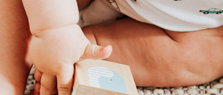 Ein Baby, das ein weißes T-Shirt mit Autoaufdruck trägt, spielt mit einem Holzblock mit blauem Waldesign auf einem Teppichboden.