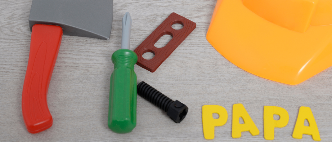 Ein gelber Schutzhelm, ein Spielzeughammer, ein Schraubendreher und ein kleines Holzstück sind auf einer Holzoberfläche angeordnet, auf der das Wort 'Papa' in Gelb steht. Über den Objekten befindet sich ein gelber Kreis mit dem Text 'EKIZ'.