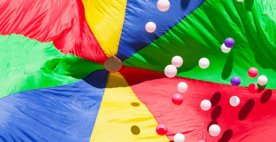 Ein farbenfroher Fallschirm mit mehreren Luftballons darauf. Der Fallschirm liegt auf einem Grasfeld.