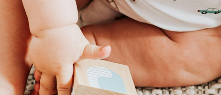 Ein Baby in einem weißen Body mit Autoaufdruck spielt mit einem Holzblock mit blauem Wal auf einem Teppichboden.