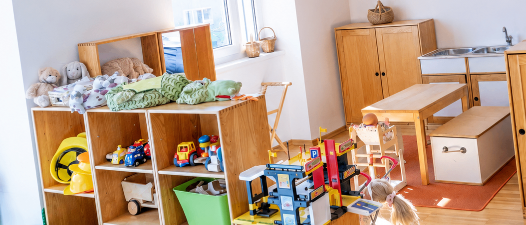 Ein Kinderspielzimmer mit Spielzeug auf Regalen, einem kleinen Tisch und Schränken. Eine orange Vorhang hängt von der Decke. Ein Waschbecken steht in der Nähe des Fensters, und ein Korb ruht auf einem Schrank.