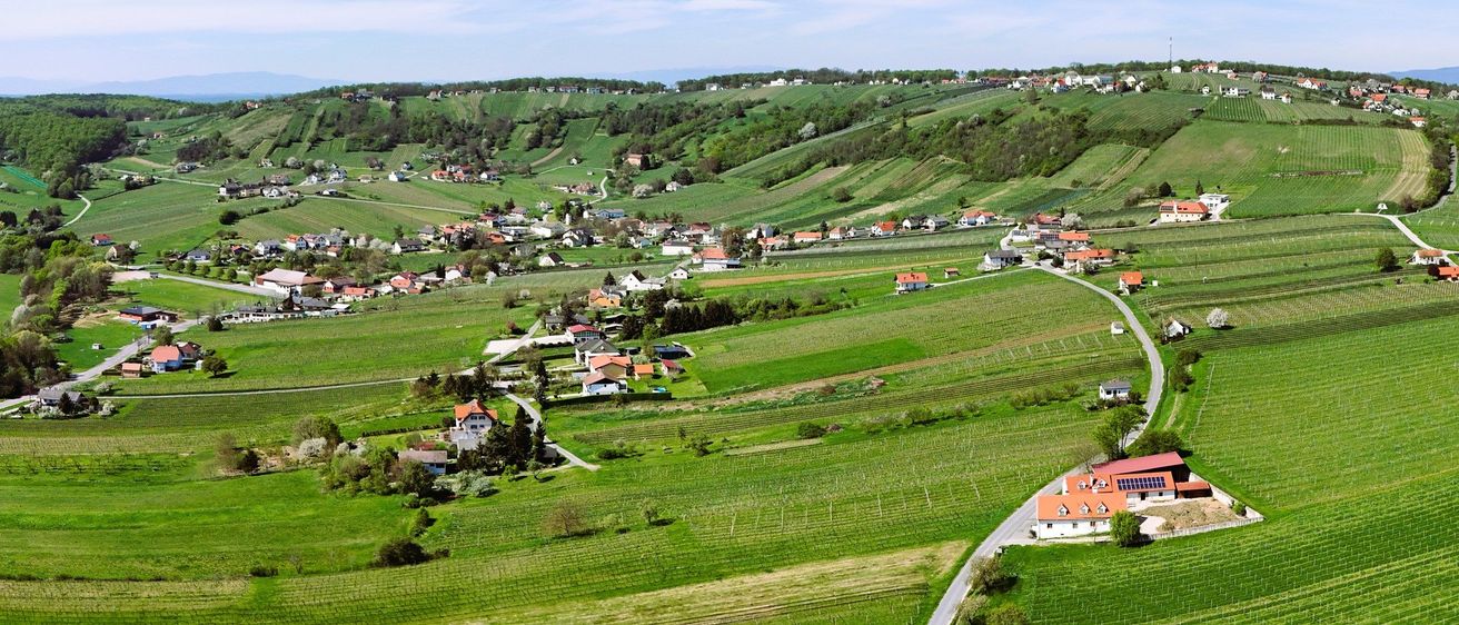 Luftaufnahme eines Hügeldorfes mit grünen Feldern und Häusern mit roten Dächern. Eine kurvenreiche Straße führt durch die Landschaft.