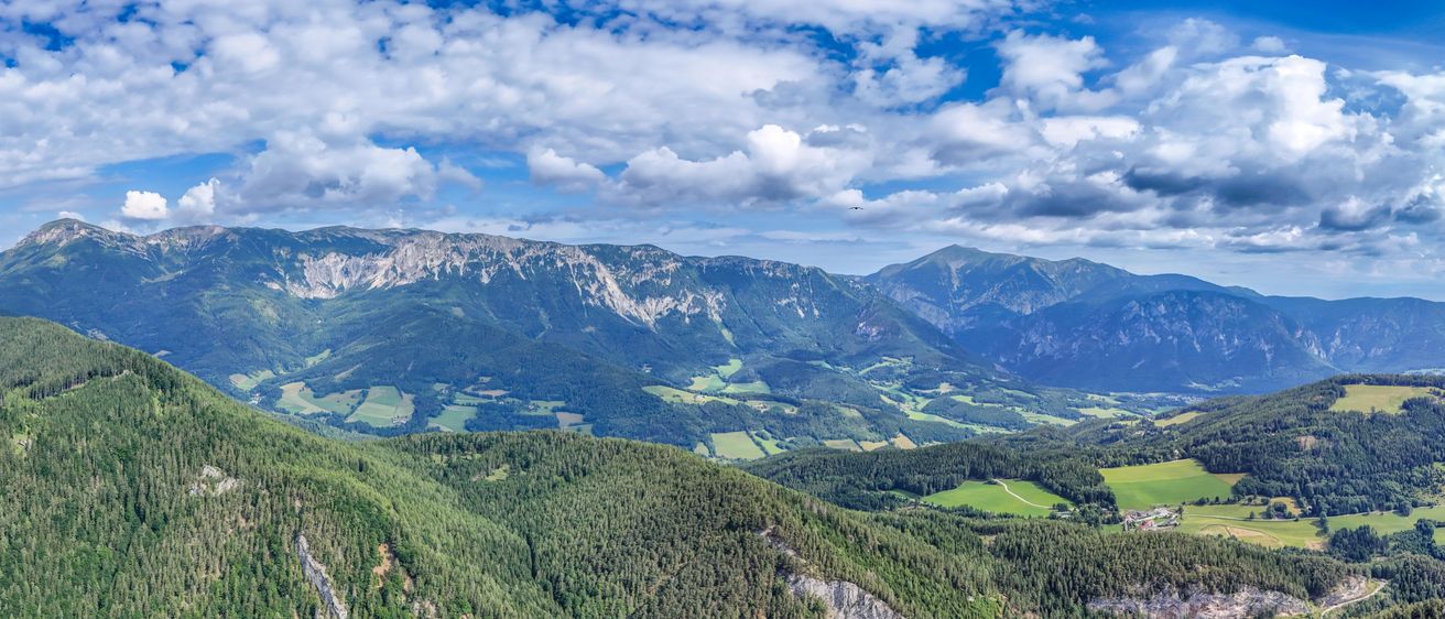 Eine hochgelegene Luftaufnahme einer Gebirgsregion mit dichten Wäldern und grünen Feldern unter einem teilweise bewölkten Himmel.