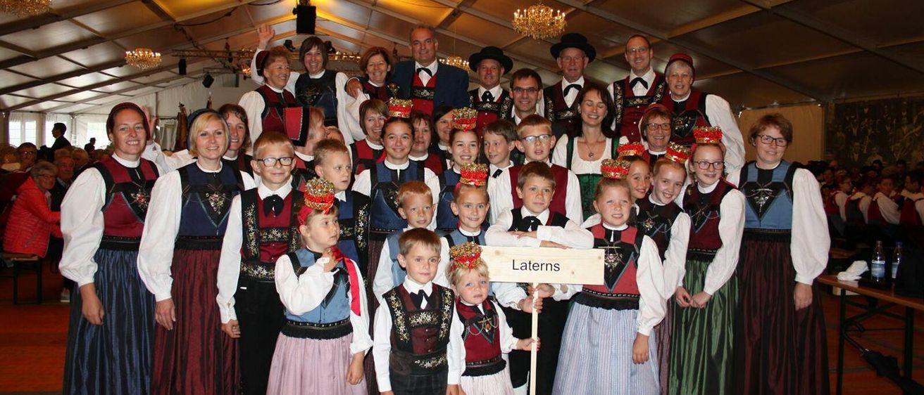 Eine Gruppe von Menschen in traditioneller Kleidung, sowohl Erwachsene als auch Kinder, stehen in einer Reihe, einige halten ein Schild mit der Aufschrift 'Laterns'. Der Ort scheint drinnen zu sein, möglicherweise für eine kulturelle Veranstaltung.