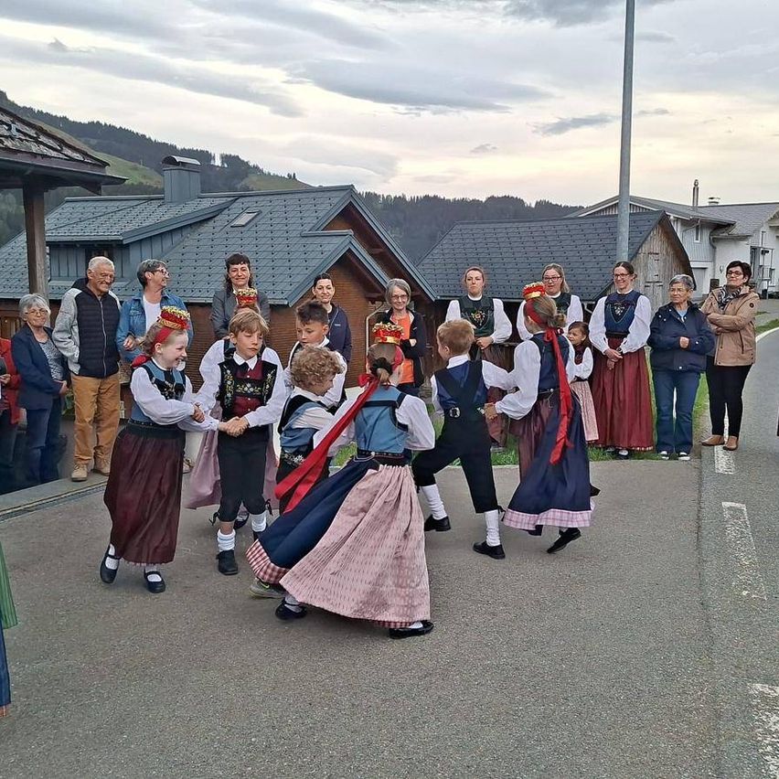 A group of children wearing traditional clothing dance in a circle, while several adults stand nearby watching. The setting appears to be an outdoor community event.