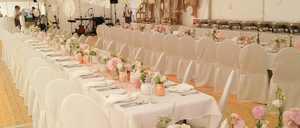 Ein großes weißes Zelt, das für eine Hochzeit vorbereitet ist, mit langen Tischen, die mit Blumensträußen geschmückt sind, und weißen Stühlen. Ein Buffetbereich ist im Hintergrund zu sehen, und verschiedene dekorative Gegenstände hängen von der Decke.