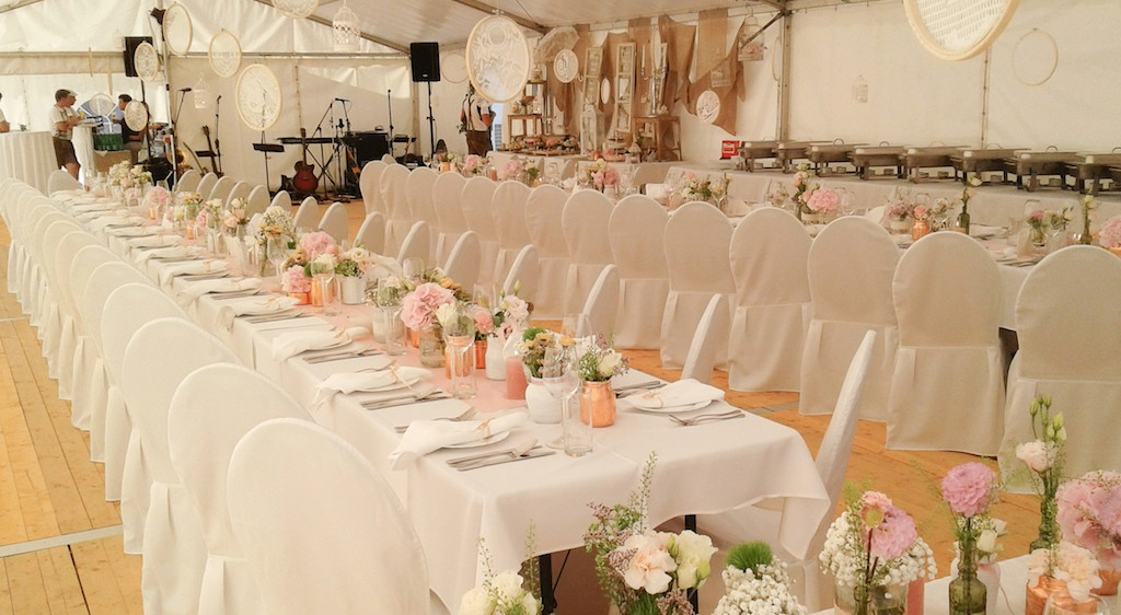 Ein großes weißes Zelt, das für eine Hochzeit vorbereitet ist, mit langen Tischen, die mit Blumensträußen geschmückt sind, und weißen Stühlen. Ein Buffetbereich ist im Hintergrund zu sehen, und verschiedene dekorative Gegenstände hängen von der Decke.