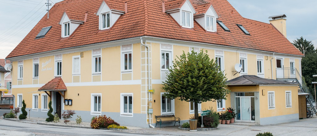 Ein gelbes Gebäude mit rotem Dach, mehreren Fenstern und einem Schornstein. Ein Baum und eine Bank befinden sich im Vorgarten.