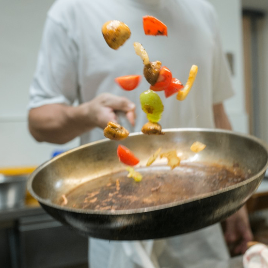 Ein Koch in einem weißen T-Shirt wirft Essen in einer Bratpfanne. Verschiedenes Gemüse und Fleisch werden in die Luft geworfen. Die Szene spielt sich in einer Küche ab.
