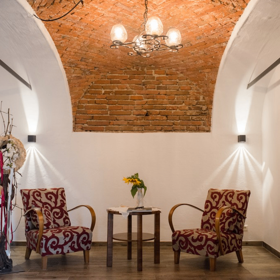 Ein Raum mit einem gemauerten gewölbten Dach, weißen Wänden und zwei rot gemusterten Stühlen. Ein kleiner Tisch hält eine Vase mit gelben Blumen, und eine Kronleuchter hängt von der Decke. Licht reflektiert an den Wänden.