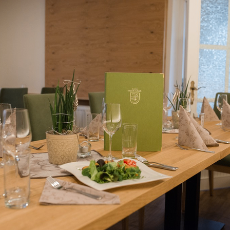 Ein für eine Mahlzeit gedeckter Esstisch mit Weingläsern, Besteck, einem grünen Menü und einem Salatteller. Der Tisch ist aus Holz und die Stühle grün.