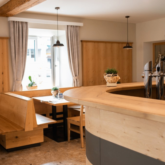 Ein Innenansicht eines Restaurants mit Holzbänken, einem runden Tisch, einer Bartheken und hängenden Lampen. Topfpflanzen und eine Uhr schmücken den Raum.