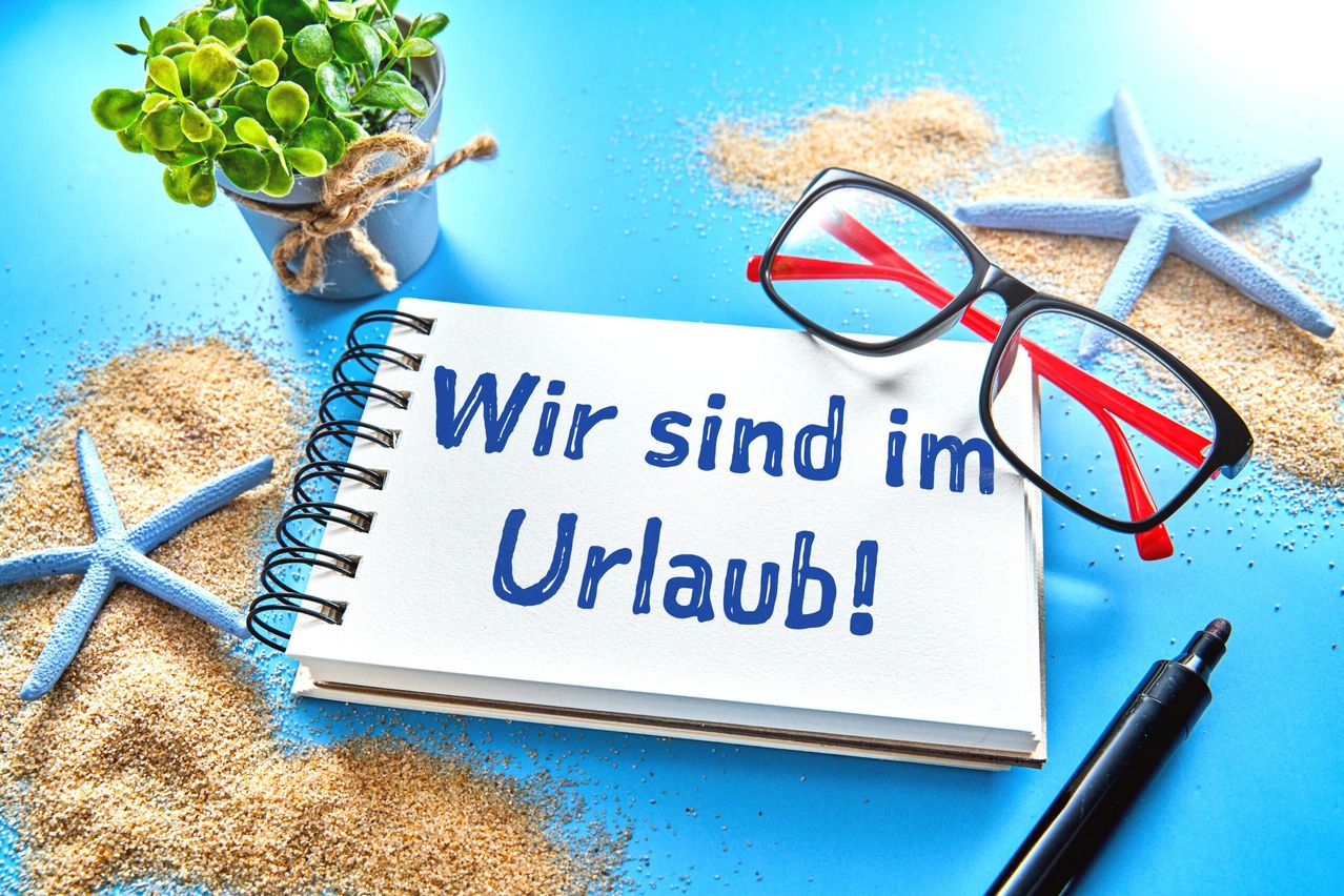 A spiral notebook with the German text 'Wir sind im Urlaub!' written in blue is placed on a blue surface with a small potted plant, a pair of glasses, and a starfish.