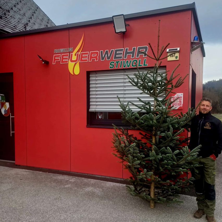 Ein Mann steht neben einem Weihnachtsbaum vor einem roten Gebäude mit der Aufschrift 'FEUERWEHR STIWOLL'.
