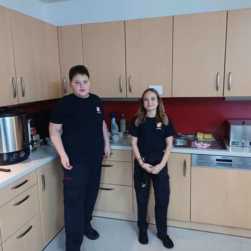 Zwei Personen in identischen schwarzen Uniformen stehen in einer Küche mit Schränken, einem Waschbecken und einer Kaffeemaschine. Einer hält einen Stift und Papier, und der andere posiert mit einem leichten Lächeln.