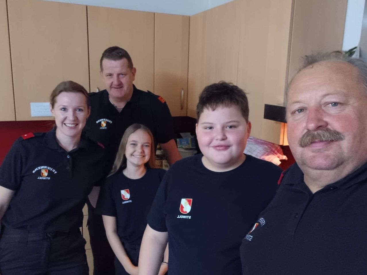 Fünf Personen posieren für ein Foto in einer Küche. Vier Erwachsene, zwei Männer und zwei Frauen, und ein junges Mädchen tragen einheitliche Kleidung. Die Küche ist gut beleuchtet und hat Holzschränke.