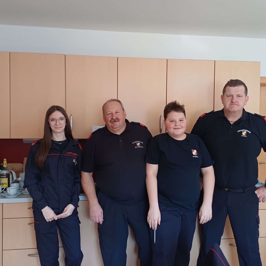 Vier Personen, wahrscheinlich Feuerwehrleute, stehen in einer Küche mit Schränken. Eine Frau und ein Junge in Uniformen und zwei Männer mit Abzeichen. Ein Mann hält einen Stift.