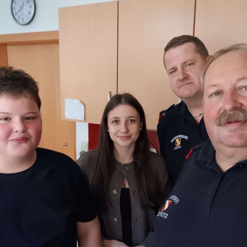 Vier Personen stehen in einem Raum. Ein Junge und eine Frau lächeln, und zwei Männer stehen hinter ihnen. Der Mann auf der rechten Seite hat einen Schnurrbart. An der Wand hängen eine Uhr und ein Blatt Papier.