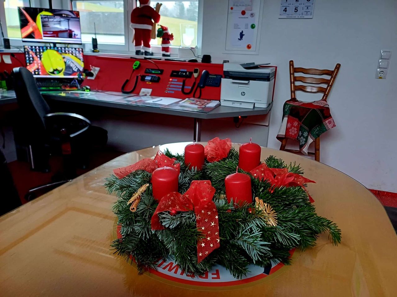 Ein festlicher Kranz mit roten Kerzen und Weihnachtsdekorationen steht auf einem Tisch. Dahinter befindet sich ein Schreibtisch mit verschiedenen Büroartikeln und ein Holzstuhl mit einer bunten Decke. Santa-Claus-Figuren sind im Fenster.