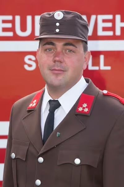 Ein Mann in einer braunen Uniform steht vor einem roten Schild. Er trägt einen Hut, eine Krawatte und ein weißes Hemd. Das Schild steht für EUIWEHL.